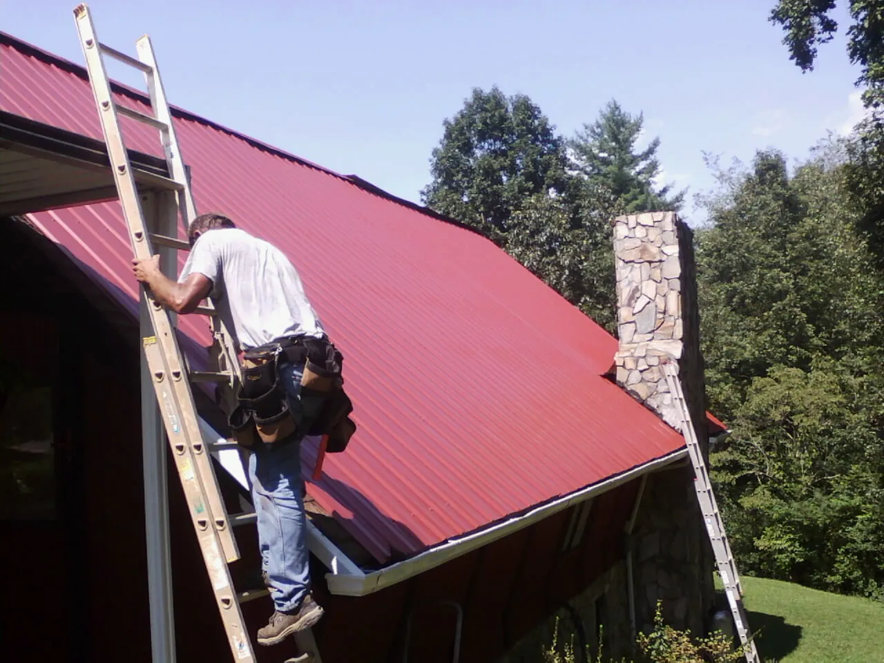 Roofer on a ladder beside a red metal roof and stone chimney — Soffit & Fascia Repair by Roofing Brothers in Yonkers